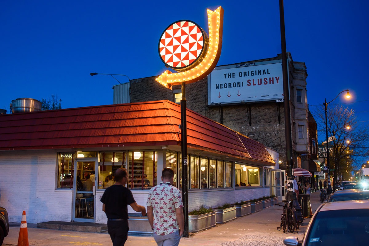 People walking by a restaurant with a neon sign at night